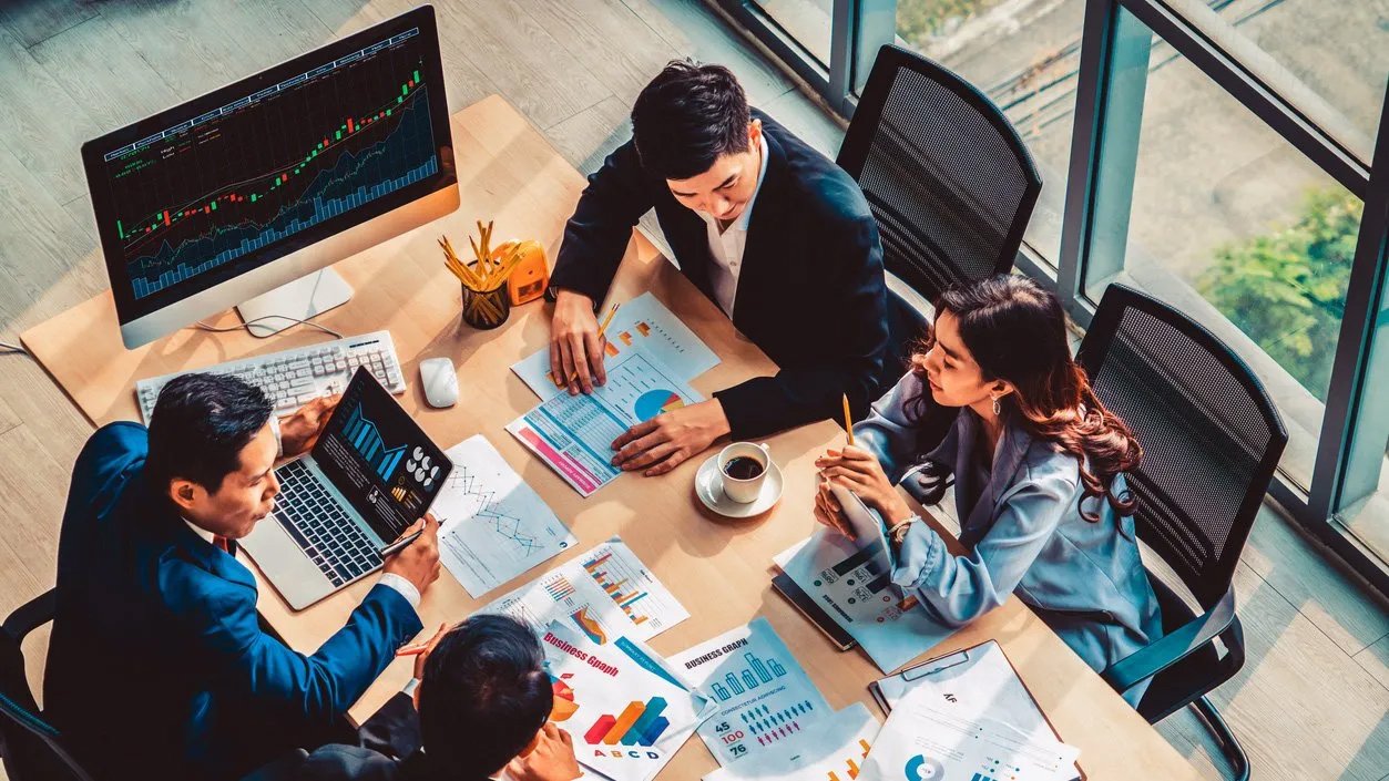 A group of business people sit at a boardroom table and discuss several graphs and charts that are displayed on papers and a computer monitor on the table.