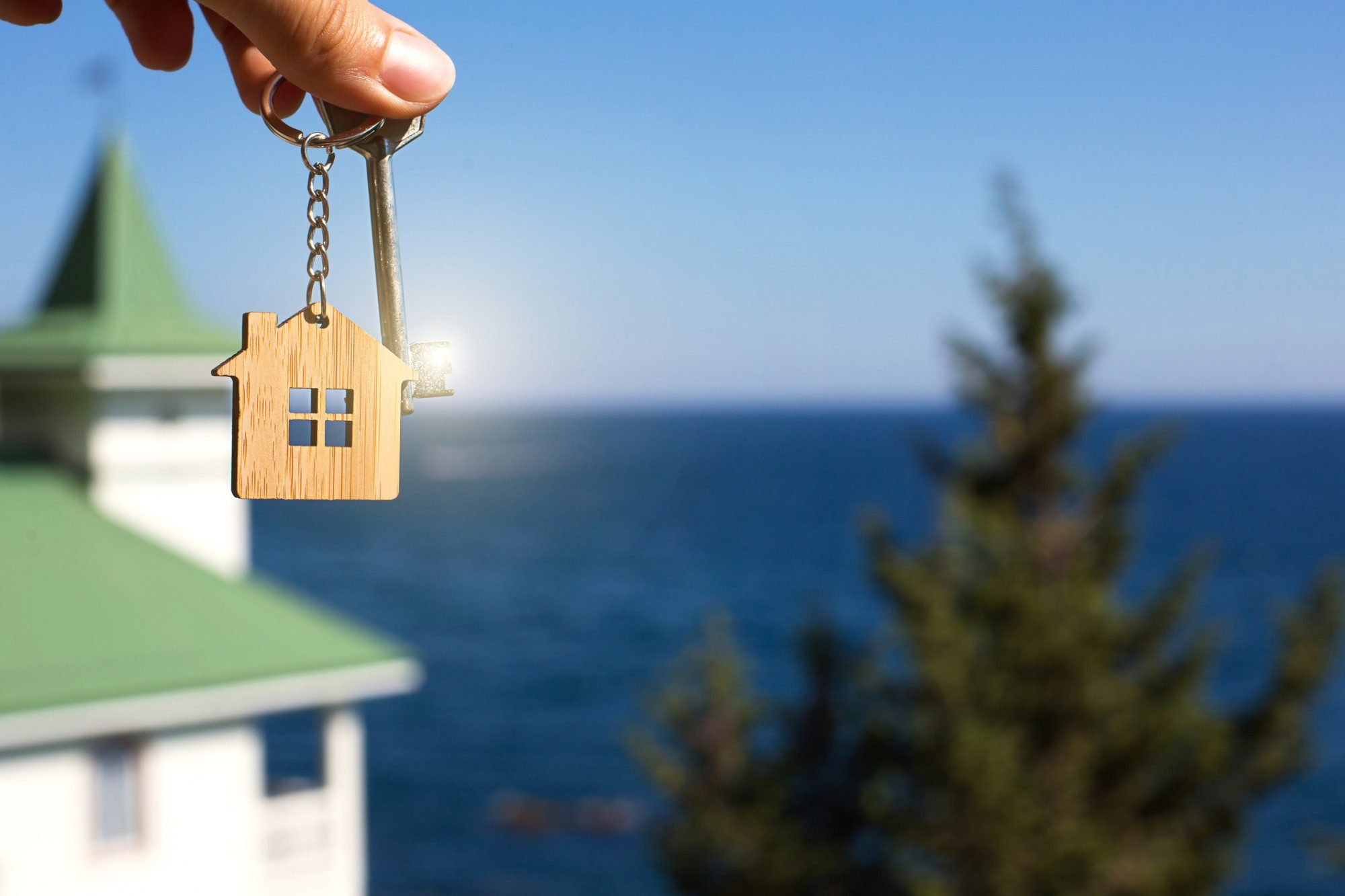 A hand holds up a keychain with a charm on it that looks like a house. There's a house in the background with an ocean view behind it.